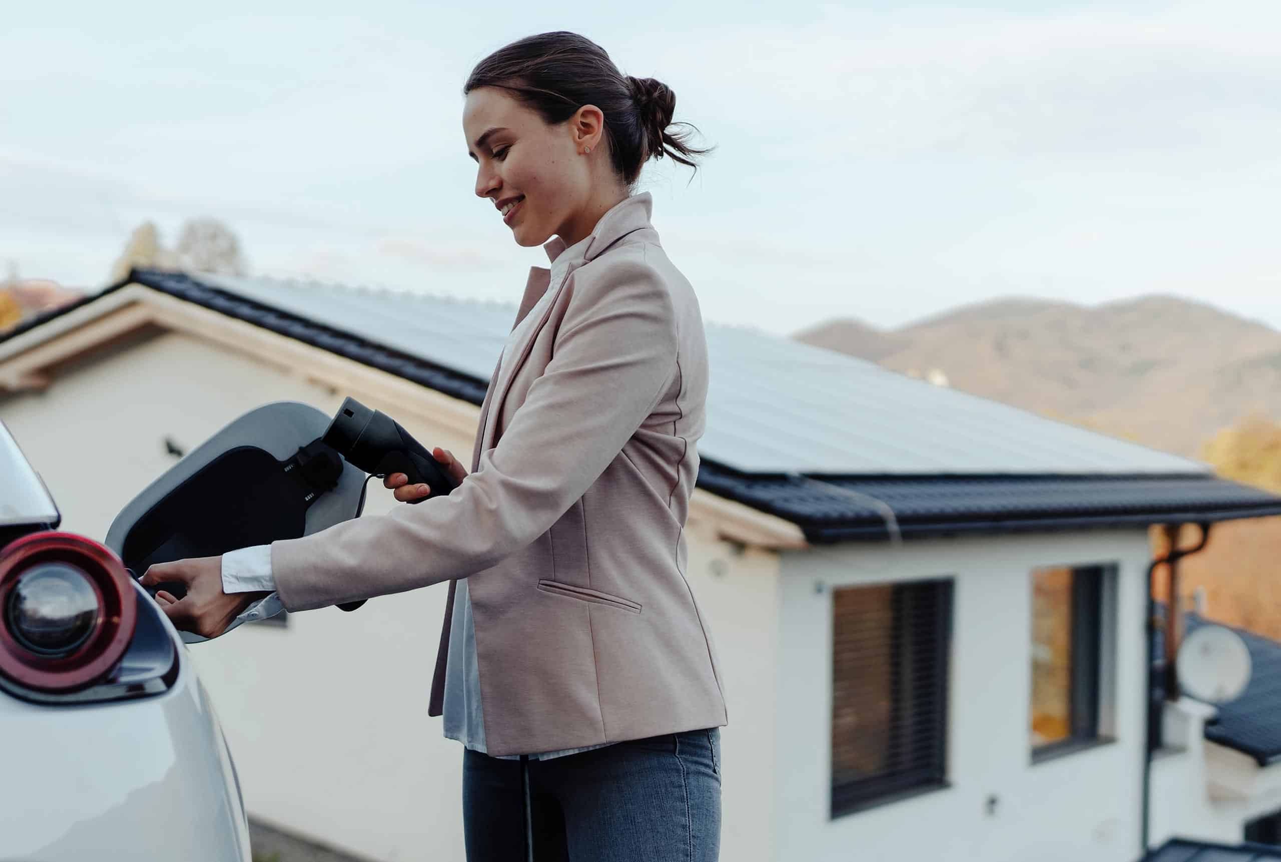 Frau lädt ihr E-Auto vor einem Einfamilienhaus mit Photovoltaikanlage auf dem Dach
